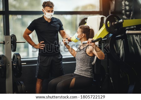 Shot of a muscular young woman with protective mask working out with personal trainer at the gym machine during Covid-19 pandemic. She is pumping up her shoulder muscule with heavy weight.