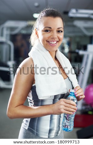 Cute Young girl standing and resting in modern gym and holding a bottle of water and towel. Looking At Camera.