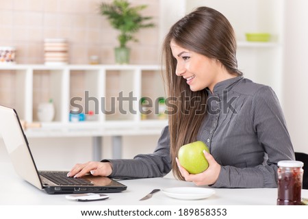 Businesswoman in the kitchen with apple reading mail on laptop before going to work.