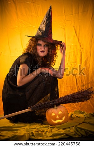 Young woman dressed like a witch. She is in dark clothing with broom and pumpkin.