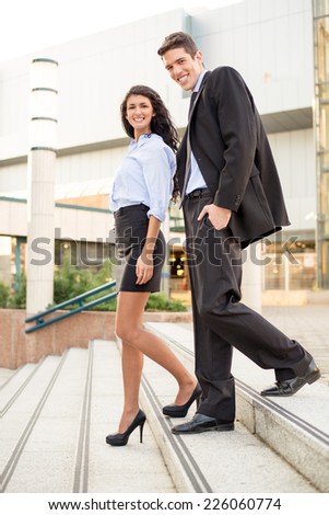 Young businessman and his colleague coming down the stairs of business building smiling looking at the camera.