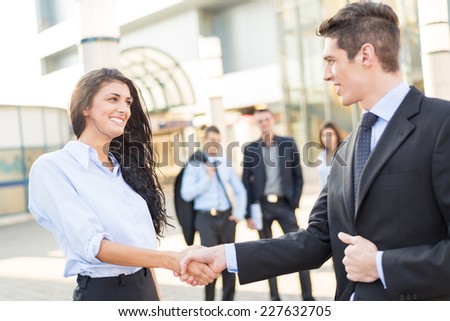 Young businesswoman and businessman shaking hands in front of the company while in the background them watching the rest of the business team.
