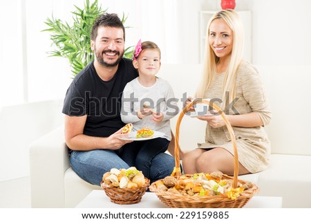Happy family in front of the table with pastry (catering). Cute little girl sitting in dad`s lap, sitting next to her mother, are holding a bun, a mother holding a cup.