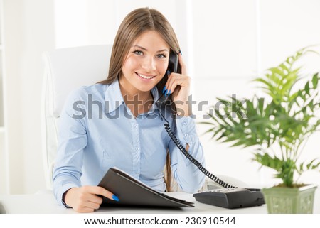 Young business woman phoning in office, sitting at an office desk with one hand holding the phone, and in another folder.