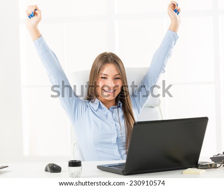 Young businesswoman sitting at an office desk in front of laptop with her hands up, looking forward to business success.