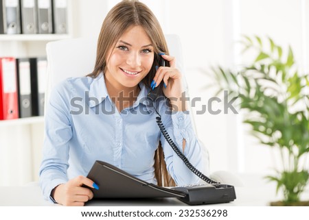 Young smiling businesswoman in her office sitting at an office desk and phoning with one hand while holding the phone, and in another folder.