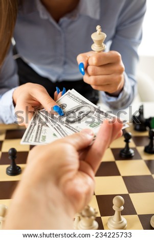 Close-up of female hands with painted fingernails over the chessboard. One hand takes money from the man\'s hand, and the other female hand holding in the hand white king chess piece.