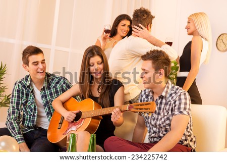 Girl playing acoustic guitar at house party, sit on the couch between two guys, and behind them is a guy who speak with two girls.