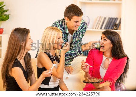 Three pretty girls sitting on the couch eating cake, a guy who stands behind them, leaning against the couch rubbing nose girl with a filling of cake.