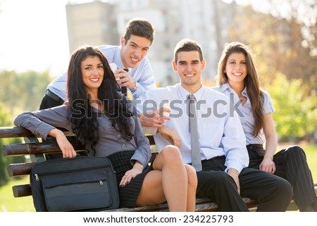 Small group of business people, elegantly dressed, on a coffee break in the park, sitting on a bench, enjoying the beautiful weather and with a smile on their faces looking at the camera.