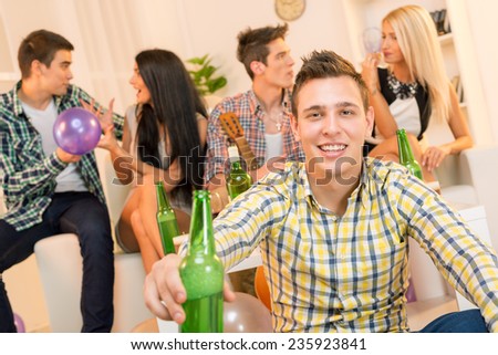 Young guy at a home party, with a smile on his face looking at the camera, holding a bottle of beer with which toasts the viewer. In the background you can see his friends sitting on the couch.