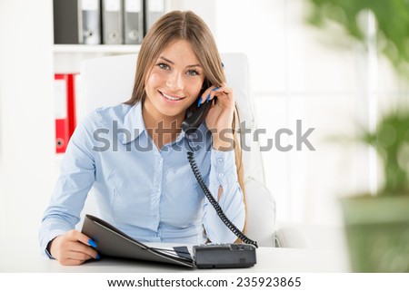 Young business woman phoning in office, sitting at an office desk with one hand holding the phone, and in another folder.