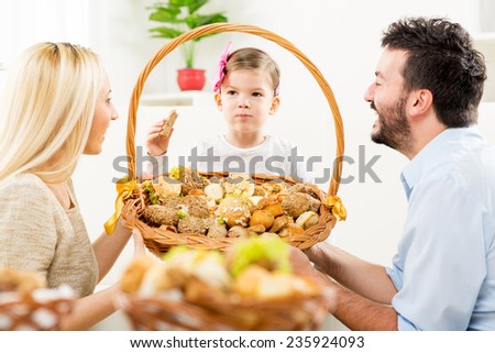 Young parents kneel in front of their little daughter with a smile watching her eat pastries from a large woven basket which together hold in their hands.