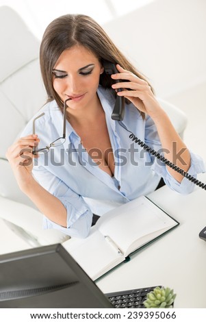 A young pretty businesswoman with cleavage sitting at a table in front of the computer and planner. While phoning, holding glasses in hand leaning on the lips.