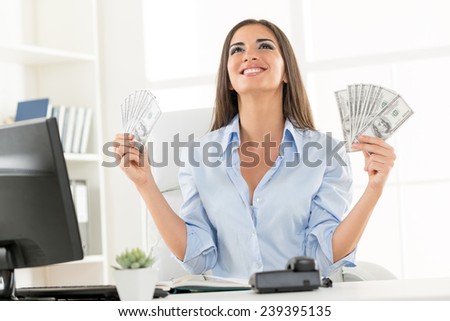 Young businesswoman in office, sitting at an office desk, holding money and with an expression of happiness on her face looking up.