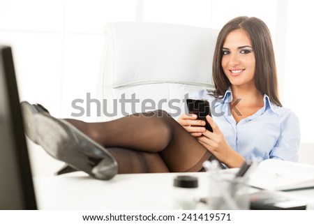 Pretty businesswoman in the office sitting on a  office chair with her feet up on the table, holding a cell phone with a smile looking at the camera.