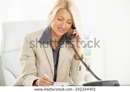 A young blonde business woman, sitting in an armchair in the office, telephoning and writing in planner, with a smile on her face.