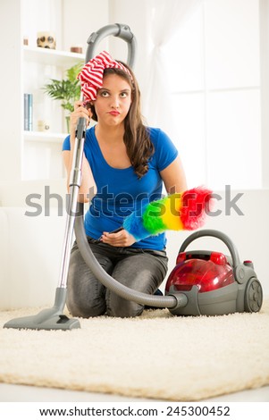A young housewife kneeling beside the vacuum cleaner with duster in hand with an expression of indecision on her face thinking where to start cleaning up the house.