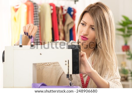 Young beautiful woman sitting in front of a sewing machine on which sets the thread, in the background you can see hanging garments.