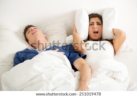 Heterosexual couple in bed, man sleeps and snoring with mouth open, while a woman irritated by snoring covers the ears with a pillow.