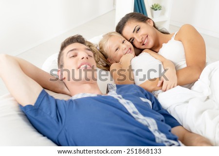 Young parents with their little daughter between them, lie in bed and with a smile looking at camera.