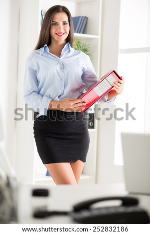Young pretty business woman in a short skirt, holding a binder and with a smile looking at the camera.