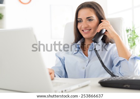 Young business woman phoning in office, sitting at an office desk in front of laptop, in which looks with a smile on her face.