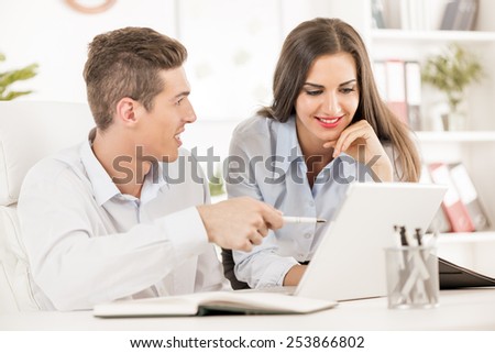 Young business people, businessman sitting at an office desk and showing something on a laptop, next to him is a businesswoman leaning against the table and looks at a laptop with a smile.