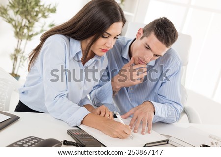 Two young business people, man and woman sitting in office at an office desk and looking at the organiser in which businesswoman writes.