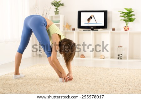 Young woman in sports clothes, athletic build, photographed from behind, doing stretching exercises in the room, in front of the TV.