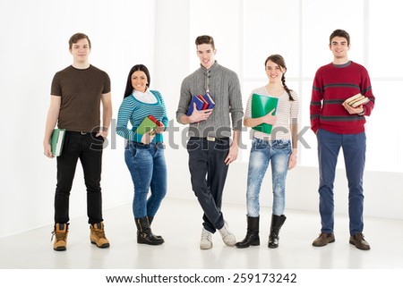 Group of cheerful students with books standing in school hall and looking at camera.