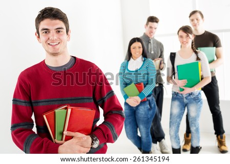 A beautiful smiling young man with books is standing in the foreground. A happy group of his friends is behind him. Looking at camera.