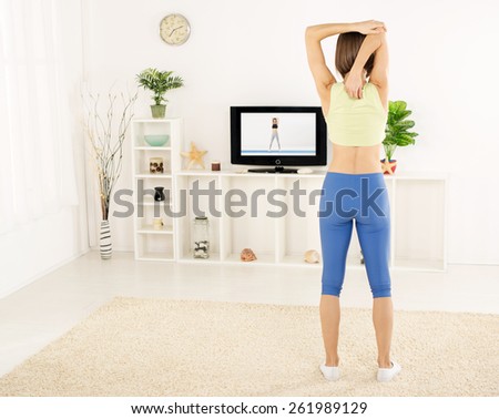 Young woman in sports clothes, athletic build, photographed from behind, doing stretching exercises in the room, in front of the TV.