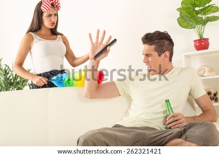 Young man sitting on sofa and watching tv. His angry wife yelling.