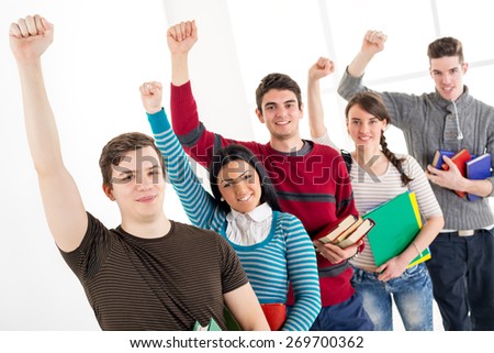 Group of cheerful students with book\'s standing In A Row with arms raised in a fist in school hall and looking at camera.