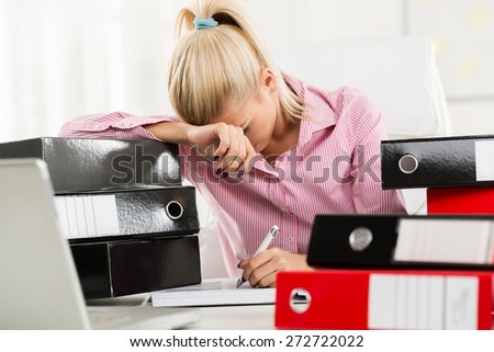 Young beautiful exhausted businesswoman sitting at a office desk between piles of binders and writes with her left hand.