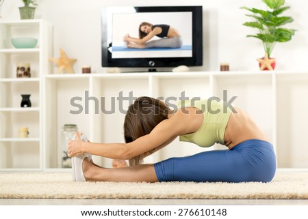 Young woman in sports clothes, doing stretching exercises in the room, in front TV.
