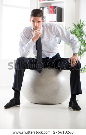 The young businessman sitting in the office on pilates ball and thinking. Looking at camera.