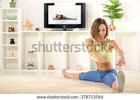 Young woman in sports clothes, doing stretching exercises in the room, in front TV.