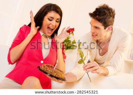 Young man with a rose in his mouth gives beautiful brunette girl birthday cake, pleasantly surprised girl with wide open mouth looking into the cake.
