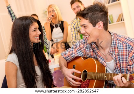 A young man with an acoustic guitar, at home party, courting a pretty girl playing to her. In the background you can see young people sitting on the couch and enjoy the atmosphere of home party.