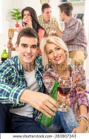 Young couple at home party with a smile on their faces, toasting with drinks that are in the foreground. In the background is they friends.