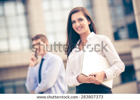 Young business partners standing in front of the building. Business woman carrying laptop while her partner use a mobile phone for business conversation.