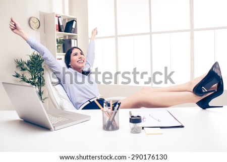 Young pretty businesswoman in the office sitting in office chair, hands up and with her feet up on a desk. In the background you can see the shelves with binders.