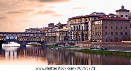 A night scene at sunset of the Ponte Vecchio bridge over the Arno River in Florence, Italy. The bridge is made of stone and houses many tourist shops, jewelers and art dealers.