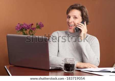 Pretty brunette working or shopping at a laptop on the kitchen table with a cup of coffee talking on the phone looking up as if interupted