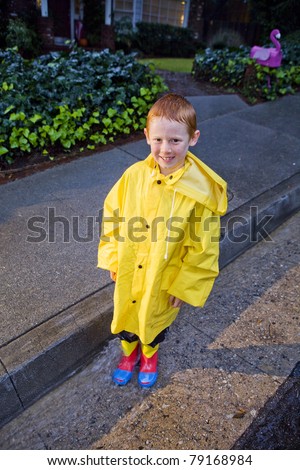 Young boy with red hair playing in the rain wearing yellow raincoat and rain boots