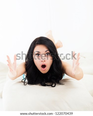 beautiful young woman surprised excited scared, terrified lying on the couch, sofa home indoors, hold hands raised palms looking at camera concept of worried, shock, fear young pretty girl