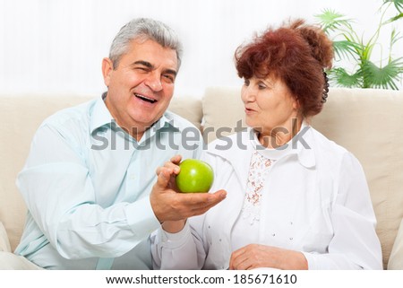 Laughing senior couple hold green apple sitting at sofa, having breakfast