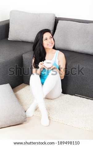 young woman hold cup of coffee think relaxing sitting on floor near couch, sofa home indoors, happy girl smile day dreaming with tea mug in hands looking up away in thought